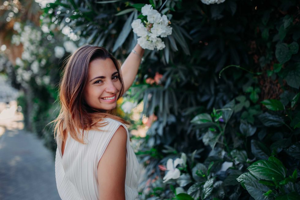 A beautiful brunette girl stands in a white dress on the background of bushes and flowers