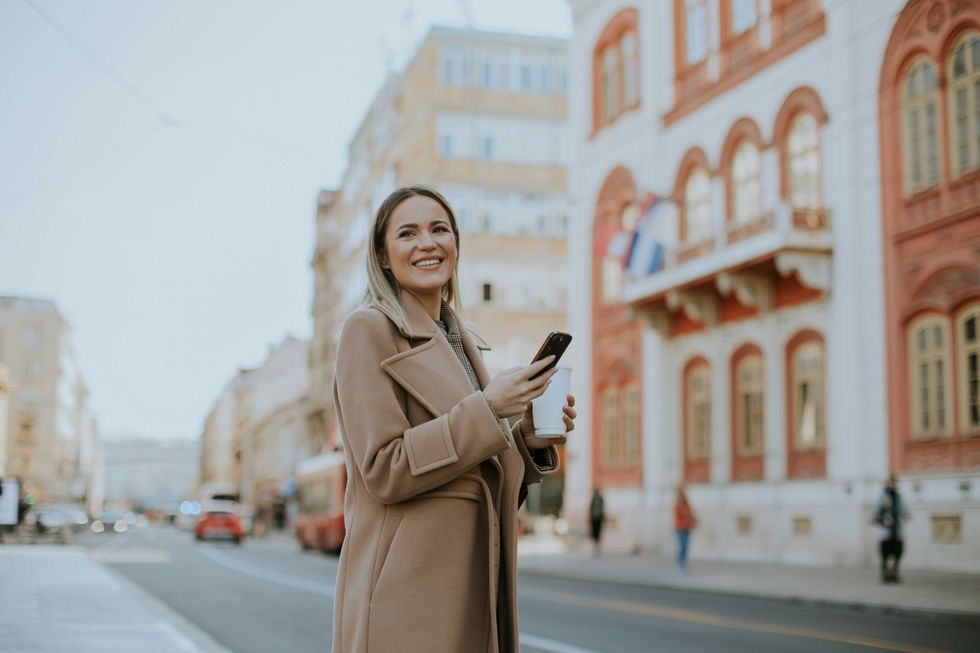 A cheerful woman stands on a city street, holding her smartphone and enjoying the sunny day. Colorful buildings line the street, creating a lively and inviting atmosphere.