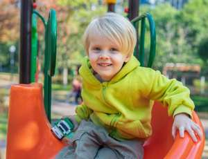 A happy blonde toddler boy sits on a bright orange slide, holding a small toy in his hand. He smiles widely in a park surrounded by colorful autumn trees