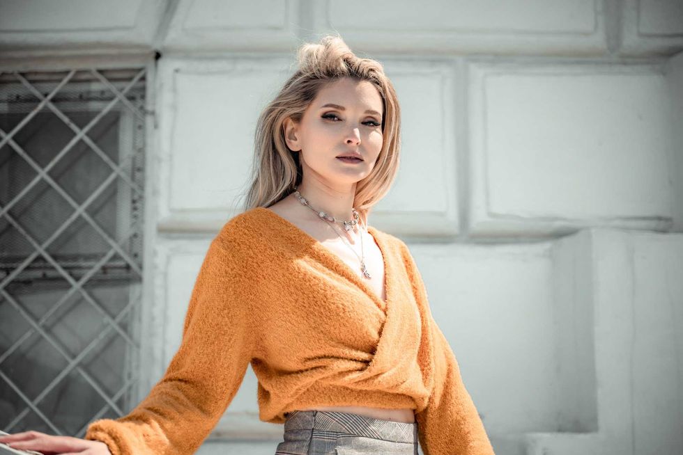 A middle-aged woman looks like a good blonde with curly beautiful hair and makeup on the background of the building. She is wearing a yellow sweater
