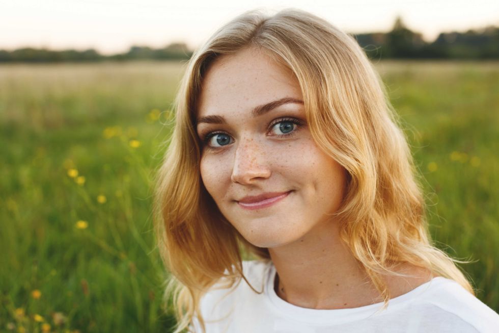 A portrait of beautiful young blue-eyed girl with light hair having charming smile and dimple on her face looking into camera standing over green background having nice time relaxing outdoor in summer