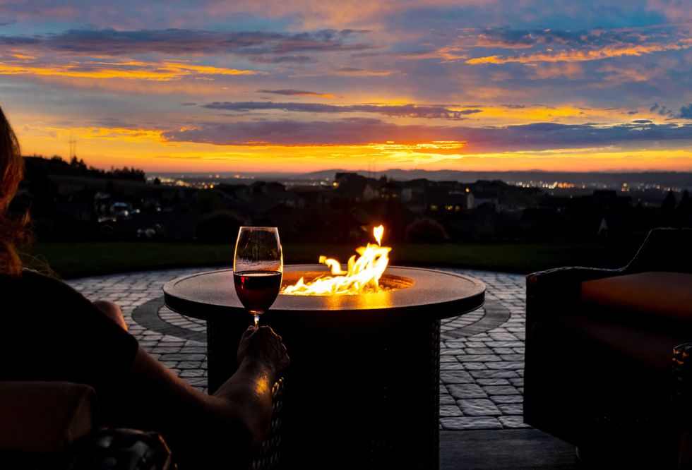 A woman drinks a glass of wine in front of a fire pit on the luxury patio of a hillside home overlooking city lights.