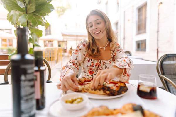 A woman sits at a table in an outdoor cafe. She holds a slice of pizza and smiles. Plates of food and drinks are on the table, and sunlight shines through the trees.