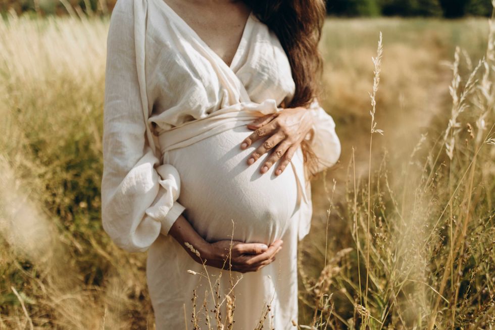 a young woman in a white linen dress wrapped her arms around her pregnant belly. body of pregnant woman in white dress closeup