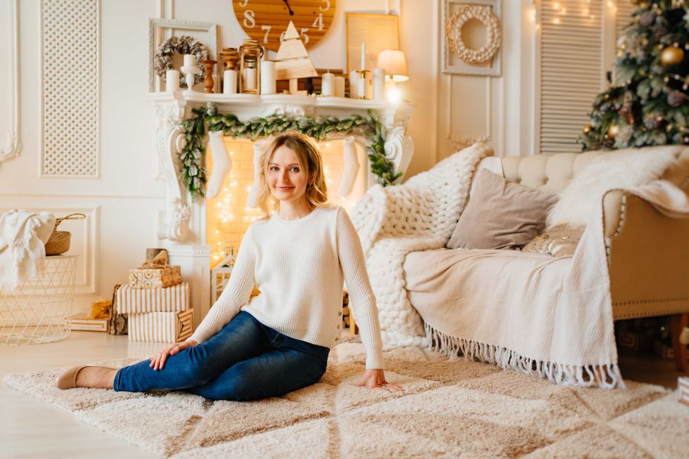 an attractive blonde woman in a white sweater on a carpet by the fireplace against the background of a Christmas tree. cozy home clothes. festive mood and decor. gift ideas.
