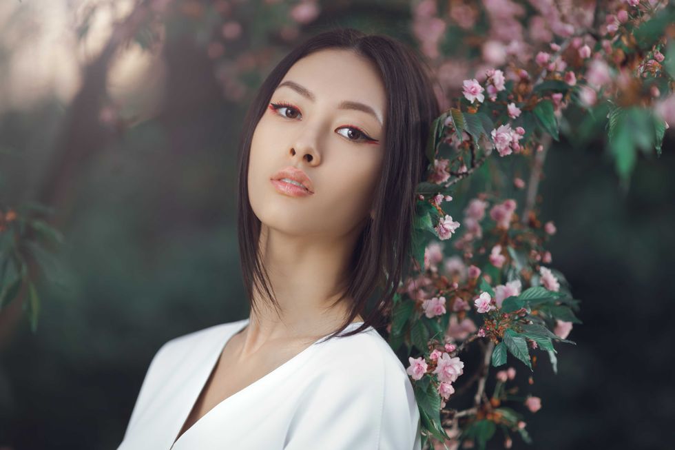 Asian woman fashion close-up portrait. Beautiful mixed race asian caucasian young girl perfect skin creartive art make-up wearing trendy white japanese kimono standing agaist flower background. Spring blossom of sakura tree in city park and elegant fashion lady.