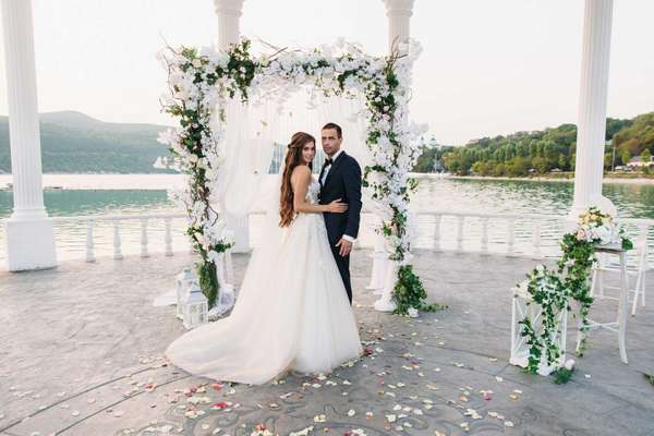Attractive groom and bride at wedding day ceremory with arch and lake on background stands together. Beautiful newlyweds, young woman in white dress and long hairs, men in black suit. Happy family concept.