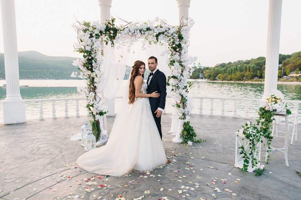 Attractive groom and bride at wedding day ceremory with arch and lake on background stands together. Beautiful newlyweds, young woman in white dress and long hairs, men in black suit. Happy family concept.