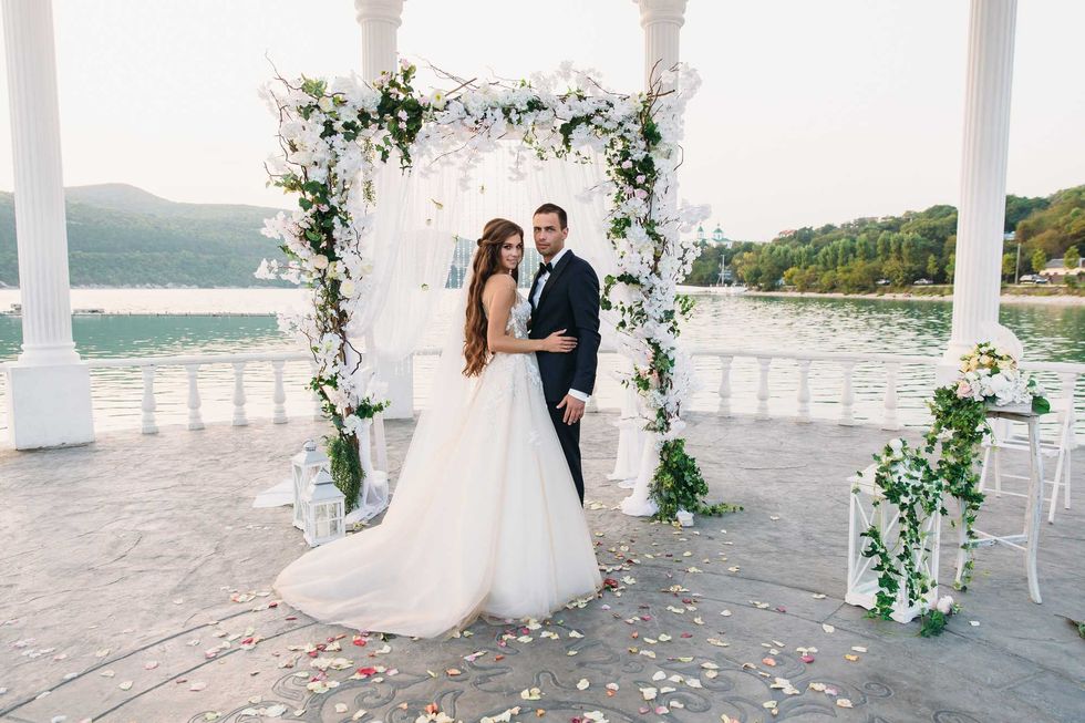 Attractive groom and bride at wedding day ceremory with arch and lake on background stands together. Beautiful newlyweds, young woman in white dress and long hairs, men in black suit. Happy family concept.