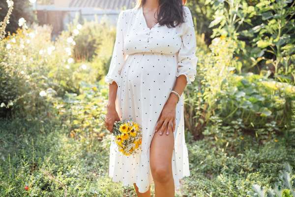 Attractive pregnant female brunette in elegant white dress standing outdoors in garden among trees and flowers. Young woman expecting baby smiles and poses in cozy green place.