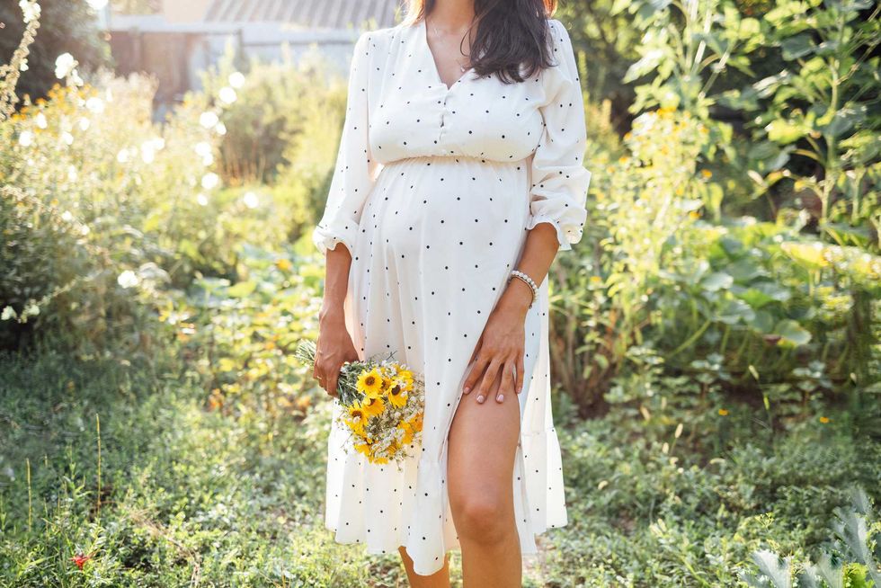 Attractive pregnant female brunette in elegant white dress standing outdoors in garden among trees and flowers. Young woman expecting baby smiles and poses in cozy green place.