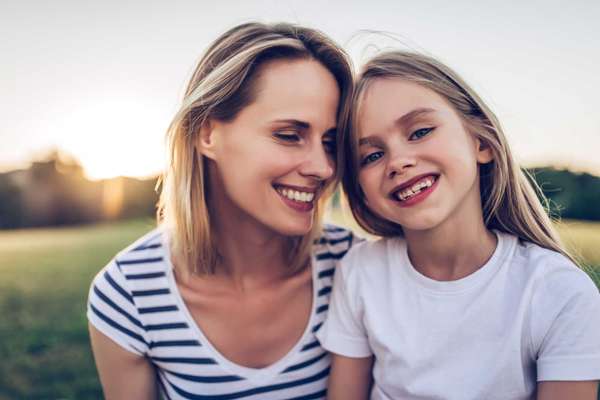 Attractive young woman with her little cute daughter are spending time together outdoors. Mom with daughter in park on a green grass during the sunset.