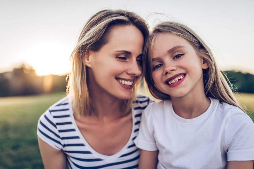 Attractive young woman with her little cute daughter are spending time together outdoors. Mom with daughter in park on a green grass during the sunset.