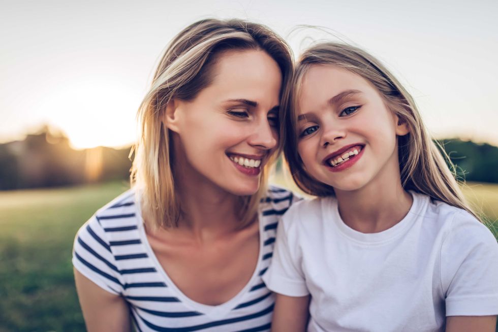 Attractive young woman with her little cute daughter are spending time together outdoors. Mom with daughter in park on a green grass during the sunset.