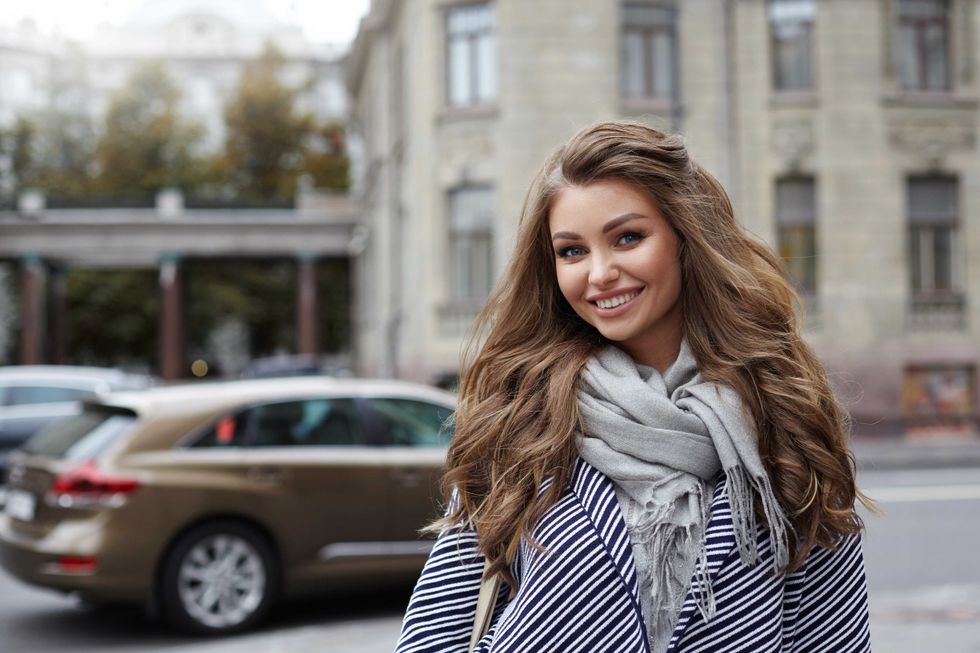 Beautiful cheerful young lady with voluminous hair wearing stylish coat and scarf posing on city street on her way to store, having joyful happy look, enjoying warm spring day. People and lifestyle