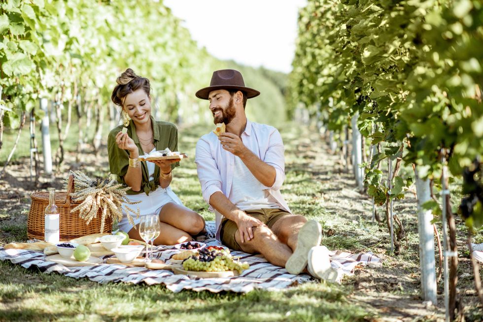 Beautiful couple having romantic breakfast with lots of tasty food and wine, sitting together on the picnic blanket at the vineyard on a sunny morning