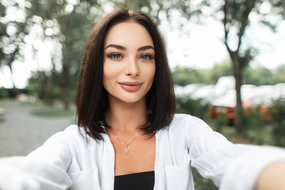 Beautiful happy woman with a smile in a fashion white shirt walking in the park and taking selfies on the phone. Female portrait of pretty girl