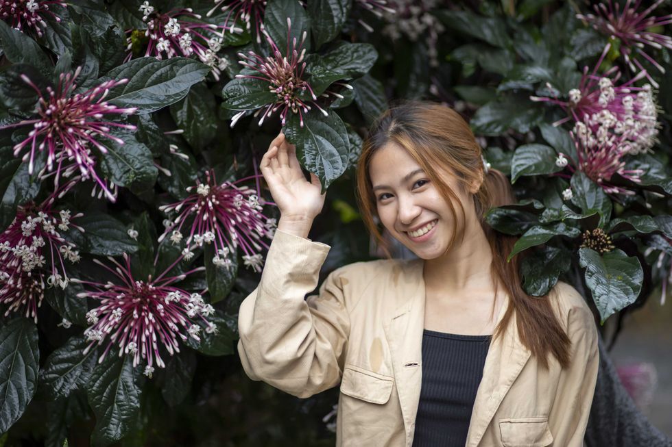 Beautiful smiling Asian woman feeling happy in an outdoor garden. She stands near natural plant with purple flowers, enjoying serene moment in nature