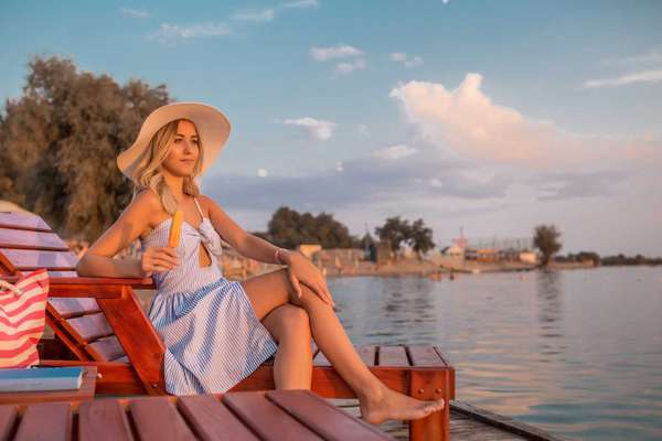 Beautiful young woman in a dress sitting on deck chair on the beach and enjoying while eating ice cream