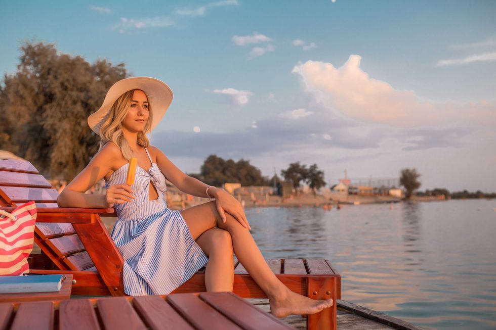 Beautiful young woman in a dress sitting on deck chair on the beach and enjoying while eating ice cream