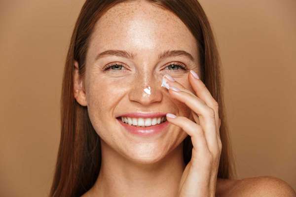 Beauty image of pretty shirtless woman smiling and applying face cream isolated over beige background
