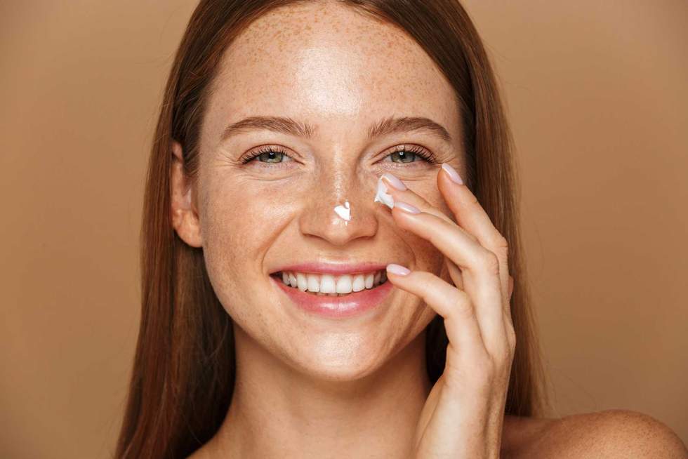 Beauty image of pretty shirtless woman smiling and applying face cream isolated over beige background
