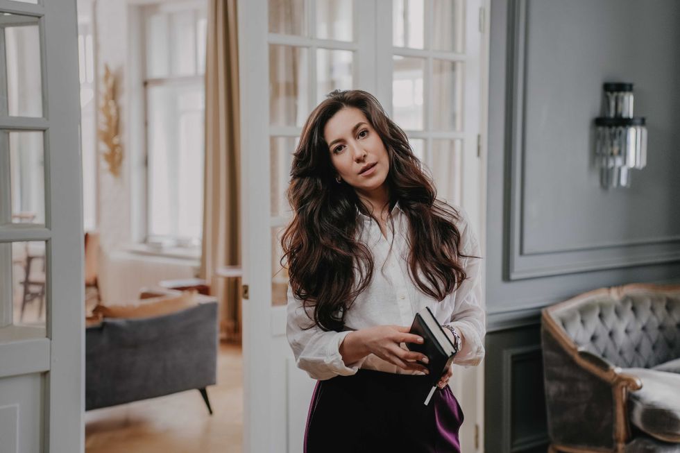 Calm brunette woman with wavy dark hair in white shirt and violet pants standing in living room, looking at camera, holding diary. Tired businesswoman at home after busy day at work. Business people.