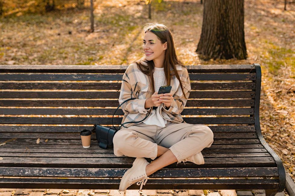 candid attractive young smiling woman sitting on bench in autumn park using phone wearing checkered coat, happy mood, fashion style trend