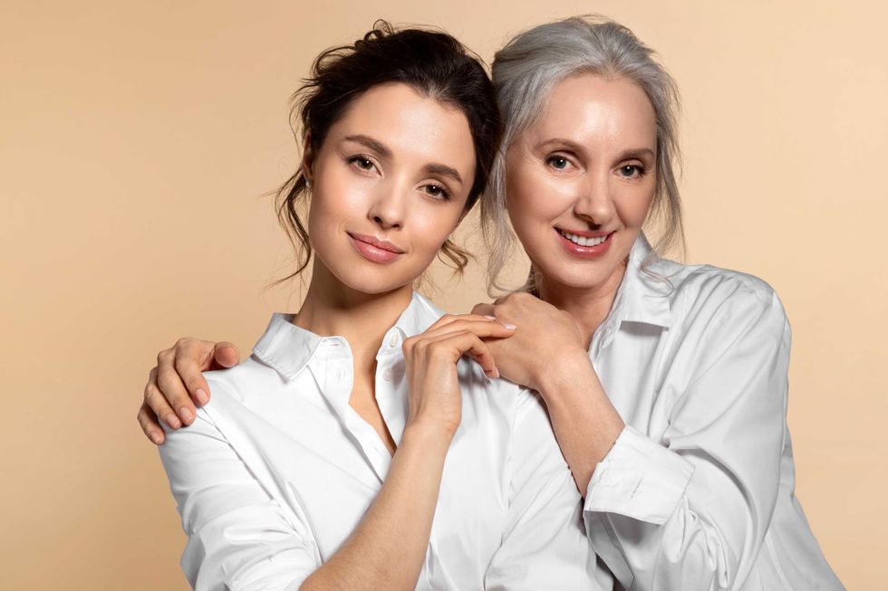 Charming beautiful mother and daughter in white casual shirt studio face forward portrait. Affectionate senior woman hugging adorable young girl shoulder from back. Adult child touching mom hand