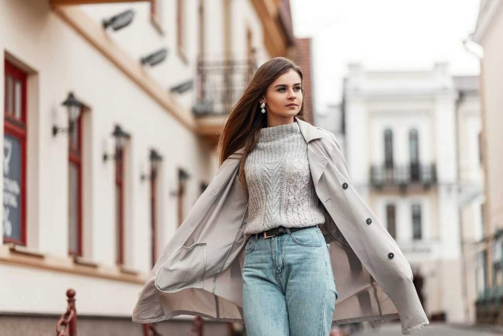 Chic modern young woman with brown hair in a stylish long trench coat in a knitted sweater in a blue jeans travels on the city. Urban girl walks on the street on a spring day.Seasonal women's clothing