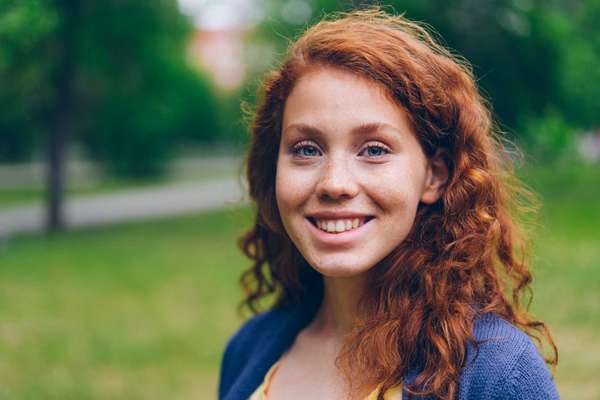 Close-up portrait of beautiful young lady with long red hair and freckles smiling looking at camera in park in summer. Nature, people and happiness concept.