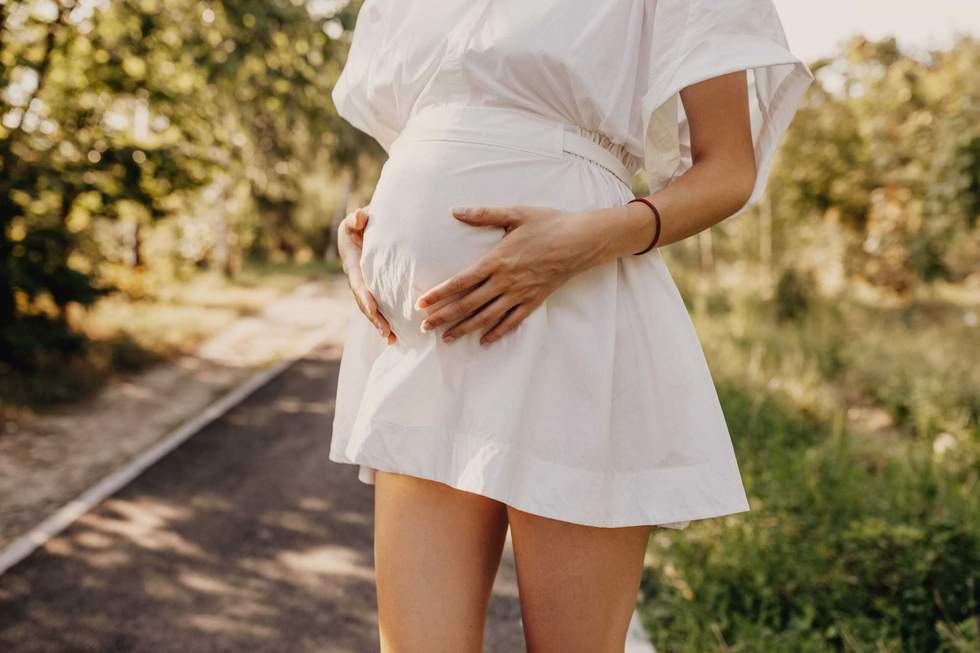 Closeup of a pregnant woman in white dress, holding hands on belly, outdoors, in a park.