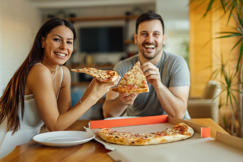 Cute couple eating pizza for breakfast, portrait. Sitting at table in pajamas and smiling at camera.