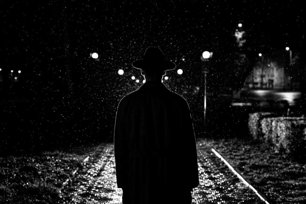 dark silhouette of a man in a hat in the rain on a night street in a city in the style of Noir