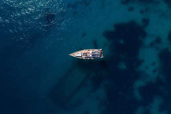 Directly above, a single sailboat rests on deep blue water. Sun glints on the sea, reefs tint patches, and the hull casts a long diagonal shadow.