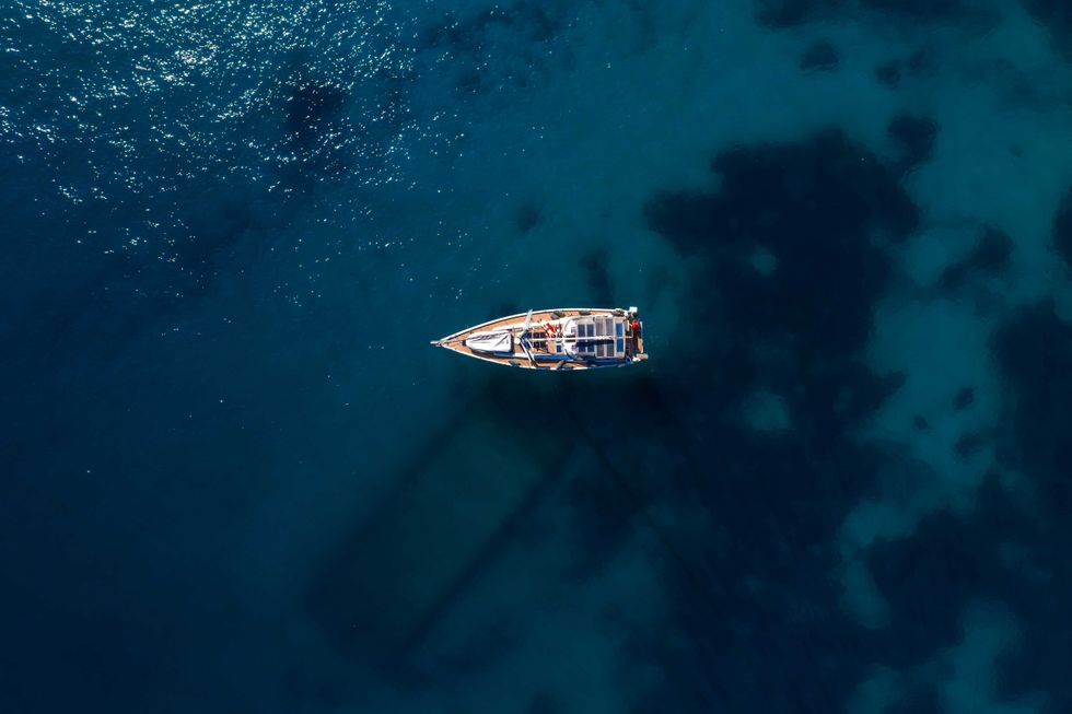 Directly above, a single sailboat rests on deep blue water. Sun glints on the sea, reefs tint patches, and the hull casts a long diagonal shadow.