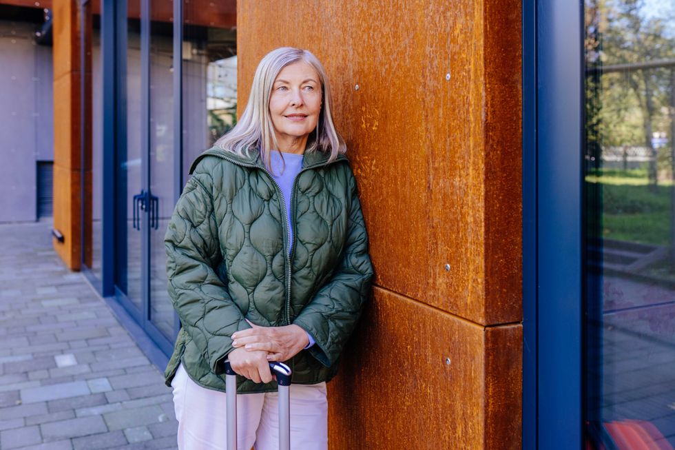 Elderly woman with long hair standing outside modern building holding suitcase relaxed in casual outfit with green jacket, suggesting travel or vacation. Adventure, leisure, and freedom in older age.