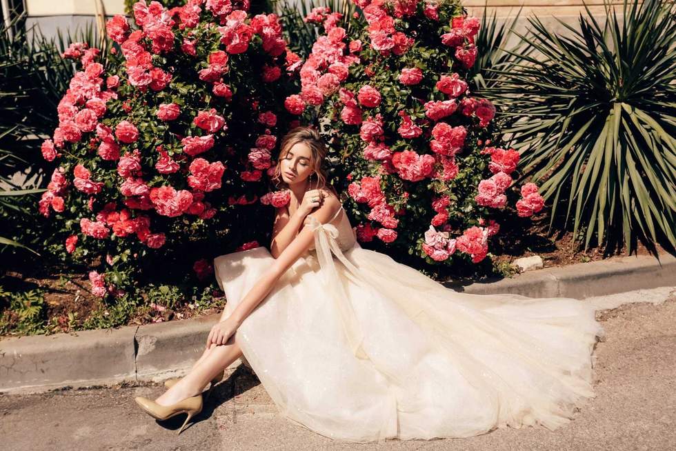 fashion outdoor photo of beautiful bride with blond hair in luxurious wedding dress posing in the spring garden with blooming roses bushes