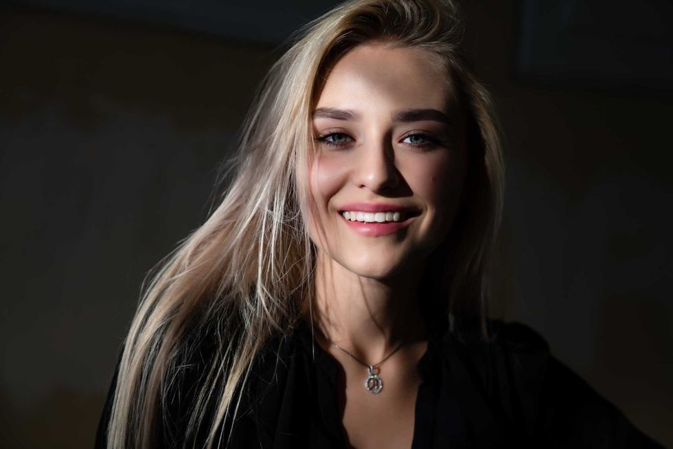 Happy smiling blonde girl with healty natural skin, dressed in black dress, stands in the shade, the rays of the sun fall on her face, outdoor photo