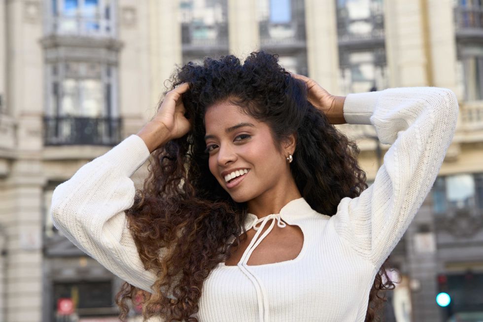 Happy young woman with beautiful curly hair smiling at camera, standing in an urban street. She is wearing a white knitted sweater