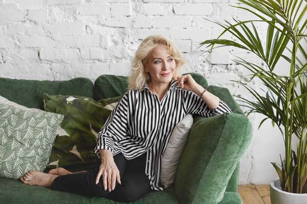 Indoor shot of beautiful middle aged European lady in elegant clothes relaxing at home, sitting barefooted on cozy couch in living room, looking sideways with positive dreamy facial expression