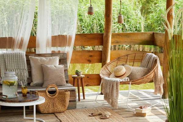 Interior design of summer gazebo by the lake with stylish rattan armchair, coffee table, sofa, pillows, plaid and elegant accessories in modern decor. Summer vibes.