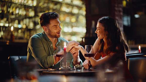 Loving boyfriend talking to girlfriend holding hands in luxury night club closeup. Smiling man making compliments to beautiful woman in red dress. Romantic couple dating in bar enjoying relationship.