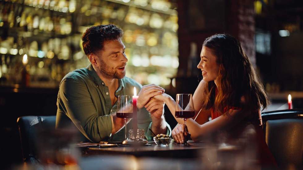 Loving boyfriend talking to girlfriend holding hands in luxury night club closeup. Smiling man making compliments to beautiful woman in red dress. Romantic couple dating in bar enjoying relationship.