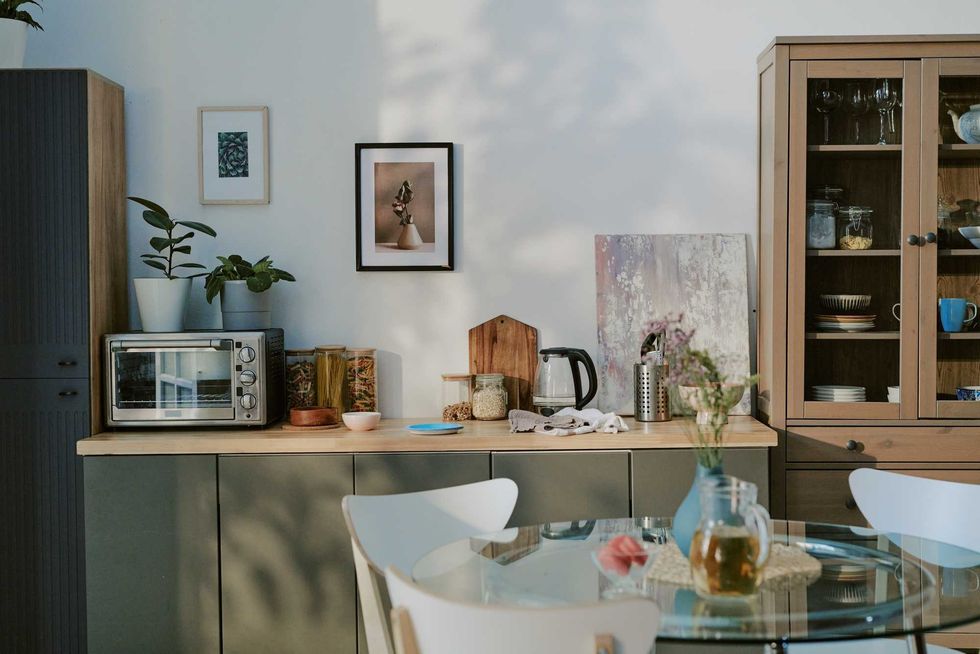 Modern kitchen interior featuring countertop with electric oven, glass jars, electric kettle, cutting board, and decorative plants, glass dining table with chairs in foreground