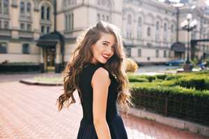Portrait from back of elefant girl with long curly hair walking on steer on old building background. She has black dress and red lips. She is smiling to camera.