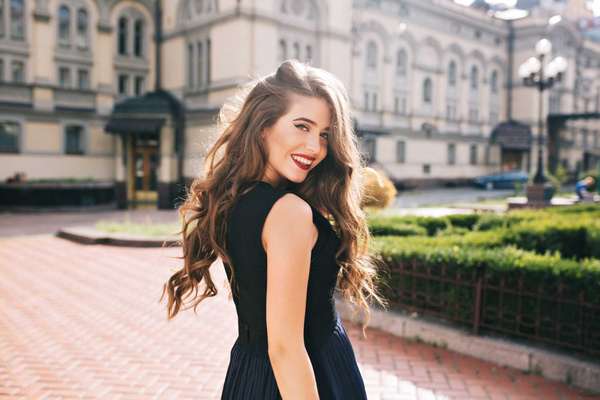 Portrait from back of elefant girl with long curly hair walking on steer on old building background. She has black dress and red lips. She is smiling to camera.