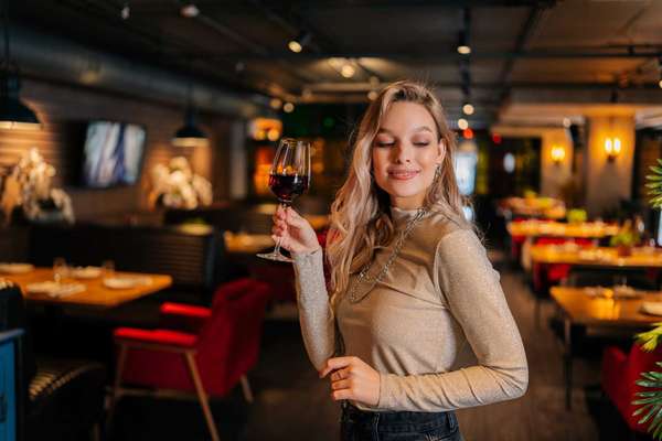 Portrait of elegant blonde woman holding glass of red wine standing in restaurant with luxury interior with dark interior, looking away. Front view of cute female posing at cafe, blurred background.
