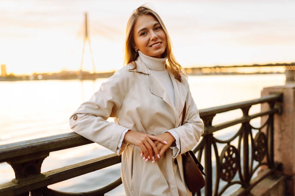 Portrait of young woman in elegant coat posing on embankment in sun rays. Beautiful woman enjoying sunset near river. Beauty, style and walks concept.