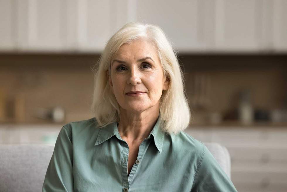 Pretty blonde old mature woman looking at camera with serious positive face expression, posing indoors in modern home apartment interior. Video conference call head shot portrait